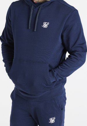 Mann trägt einen dunkelblauen Hoodie und passende Jogginghose, beide mit weißem gesticktem Logo auf Brust und Oberschenkel, Hände in der Kapuzen-Tasche.