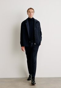 Άνδρας που προχωρά φορώντας μαύρο σακάκι, μαύρο ζιβάγκο, μαύρο παντελόνι και μαύρα παπούτσια σε απλό ανοιχτό φόντο.