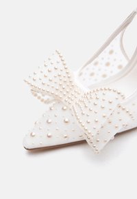 Valged teravad madalad kingad, mis on kaunistatud väikeste pärlhelmestega ja suure dekoratiivse pärlitega kaetud lipsuga.