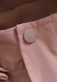 Close-up van lichtroze stof met een visgraatstructuur, met een lichtroze knoop waarop de merknaam is gegraveerd. Eenvoudige stikseldetails zichtbaar.