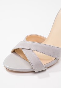 Sandale à talon haut en suède gris avec des lanières croisées, un devant lisse et un talon fin. Design minimaliste avec une texture douce et une couleur neutre.