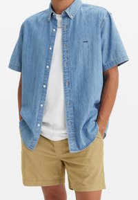 Hombre con camisa de mezclilla de manga corta azul claro sobre camiseta blanca y pantalones cortos color canela, con las manos en los bolsillos, sobre fondo blanco liso.