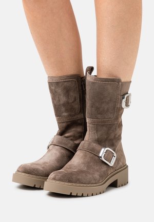 Botines de ante marrón con suela plana y robusta, que cuentan con una cremallera lateral y hebillas decorativas en las correas.