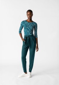 Teal långärmad topp med vitt abstrakt mönster, kombinerad med högmidjade gröna byxor med knyt framtill och fickor, bärs med vita sneakers.