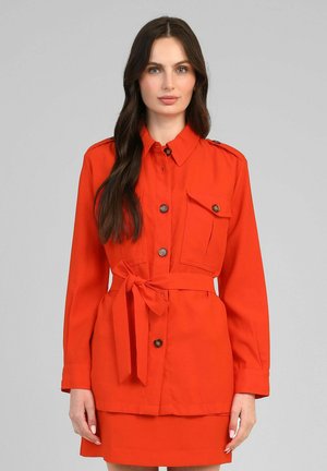 Femme aux longs cheveux foncés portant une veste ceinturée orange vif avec des boutons et une poche avant à rabat, debout devant un fond gris uni.
