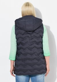 Dunkelblaues aufgeblasenes Weste mit Kapuze, ausgestattet mit einem gesteppten Zickzackmuster, kombiniert mit einem hellgrünen Dreiviertelarm-Shirt und blauen Jeans.