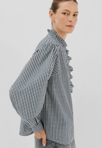 Vrouw in een zwart-witte ruitblouse met een geraffineerde voorkant en volumineuze mouwen, staat met één hand in de zak, neutrale achtergrond.