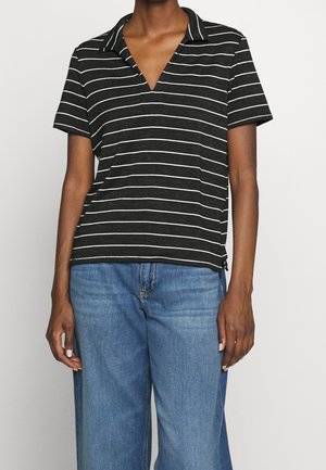 Personne portant un polo noir à manches courtes avec des rayures horizontales blanches et un jean en denim bleu, debout devant un fond uni.