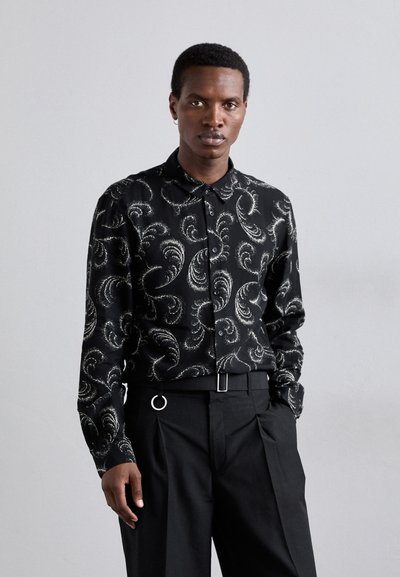 Schwarzes Hemd mit Knopfleiste, das ein weißes Paisley-Muster aufweist, lange Ärmel und einen klassischen Kragen hat. Trägt dazu taillierte schwarze Hosen mit Gürtelschlaufen.