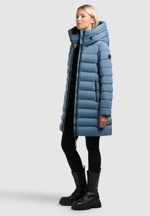 Light blue puffer coat with a hood, featuring quilted sections, zippers, and side pockets, worn over black leggings and combat boots.
