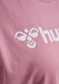 Zbliżenie na różową tkaninę z białym geometrycznym wzorem pszczoły oraz częściowym białym tekstem na górnej części klatki piersiowej ubrania.