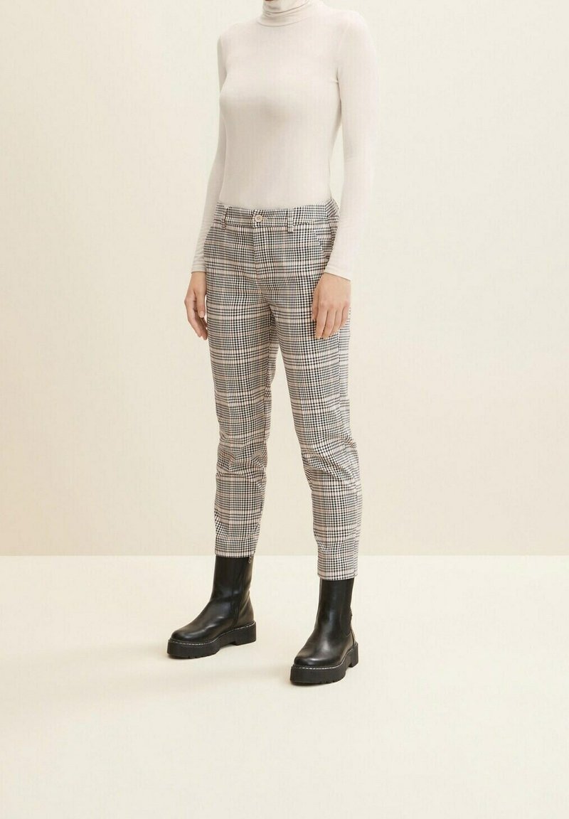 Halsen är hög på en beige topp som bärs med rutiga grå byxor, som har en smal passform, och svarta ankelboots med tjocka sulor.