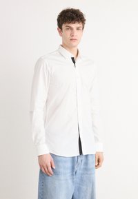 Camisa blanca de algodón de manga larga con cuello abotonado, presenta una tapeta frontal y un corte ajustado, combinada con jeans de mezclilla azul.