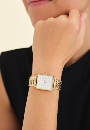 Montre-bracelet dorée avec un cadran carré, un cadran blanc, des index dorés et un bracelet en maille métallique. L'affichage indique la date ; portée au poignet d'une personne.