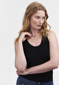 Young woman with long red hair wears black sleeveless top and blue jeans, holding a gold necklace with a red pendant near her neck.