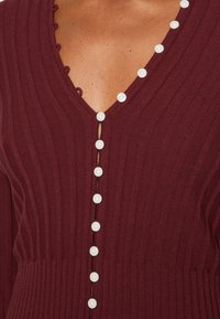 Viininpunainen neulepaita, jossa on ribbineulottu pinta, V-kaula-aukko ja valkoiset nappikoristeet keskellä. Yksityiskohtainen aaltoileva kaula-aukko.