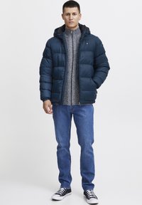 Tamsiai mėlynas pūkinis švarkas su kapisonu, užtrauktuku ir elastingais rankogaliais, dėvimas ant tekstūruoto pilko megztinio, derinamas su šviesiai mėlynomis džinsais ir juodais sportbačiais.