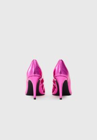 Et par skinnende metallisk pink højhælede sko vist fra bagsiden mod en ensfarvet hvid baggrund.