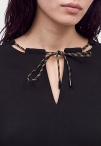 Gros plan d'une personne portant un haut noir avec un cordon à motif vert, blanc et rouge noué en un nœud papillon au niveau de l'encolure.