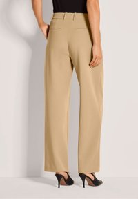 Persoon in beige hoogwaardige wijde broek en zwarte hoge slingback schoenen, staand tegen een effen achtergrond.