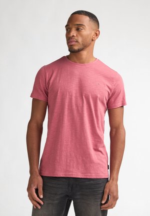 Kurzärmeliges T-Shirt mit Rundhalsausschnitt aus meliertem rosafarbenem Stoff, mit einem dezenten Logo auf der Brust, kombiniert mit dunklen Jeans.