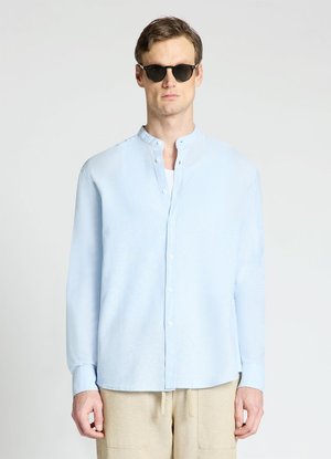 Uomo che indossa occhiali da sole neri, camicia azzurro chiaro con collo alla coreana e pantaloni beige, in piedi davanti a uno sfondo chiaro e uniforme.