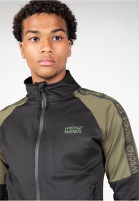 Chaqueta de cremallera negra y verde olivo con cuello alto, logo "WRONG FRIENDS" en el pecho y la manga, tejido texturizado, diseño ajustado.