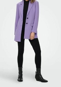 Laventeli bleiseri, jossa on viistetty kaulus, yksirivinen kahdella napilla, yhdistetty mustaan puseroon ja kapeisiin mustiin housuihin. Mustat nilkkurit.