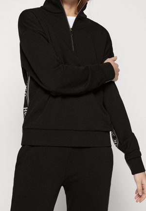 Person trägt schwarzen Sweatshirt mit Reißverschluss und weißen Textstreifen an den Ärmeln, kombiniert mit passenden schwarzen Hosen, steht vor einfarbigem Hintergrund.