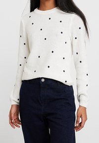 Weißer, langärmeliger Strickpullover mit verteilten schwarzen Punkten, kombiniert mit hochgeschnittenen dunkelblauen Jeans.