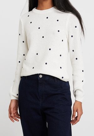 Hvid langærmet strikket sweater med spredte sorte prikker, båret med højtaljede mørkeblå jeans.