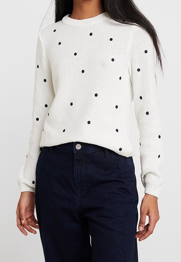 Weißer, langärmeliger Strickpullover mit verteilten schwarzen Punkten, kombiniert mit hochgeschnittenen dunkelblauen Jeans.