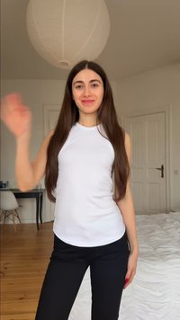 Νεαρή γυναίκα με μακριά καστανά μαλλιά, φορώντας λευκό αμάνικο τοπ και μαύρο παντελόνι, χαιρετάει σε ένα υπνοδωμάτιο με ξύλινο πάτωμα και λευκούς τοίχους.