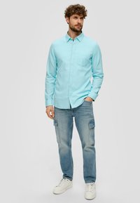 Helles blaues Hemd mit langen Ärmeln und einer Brusttasche, kombiniert mit hellblauen Cargo-Jeans und weißen Turnschuhen.