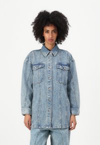 Lys blå denimjakke med spiss krage, to brystlommer og knapping foran. Har en løs passform og synlige sømdetaljer.