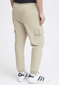 Beige cargobroek van zachte stof, met elastische boorden, zijzakken en een achterzak met een label. Gdraagt met witte sneakers.