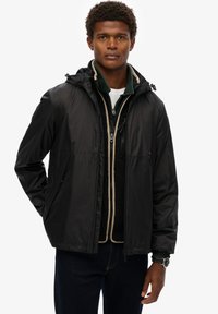 Zwarte lichtgewicht jas met een capuchon, voorzien van ritszakken, elastische manchetten en een soepele textuur. Gelaagd over een groene fleece.