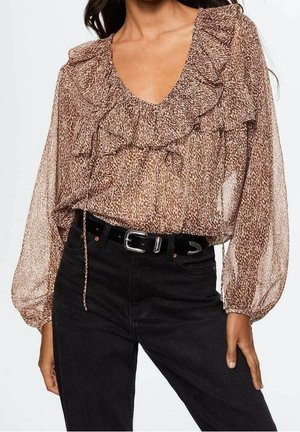 Kobieta ubrana w brązową bluzkę w wzory z falbanami i przezroczystymi bufiastymi rękawami, włożoną w czarne dżinsy z wysokim stanem i czarnym paskiem.