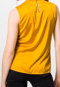 Gelbes ärmelloses Oberteil mit glatter Textur, das ein Schlüsselverschluss-Design auf der Rückseite und eine lässige Passform aufweist, die elegant fällt.