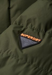 Tela acolchada de color verde oliva con textura de acolchado; presenta un parche triangular negro con "SUPERDRY" en un patrón naranja y gris.