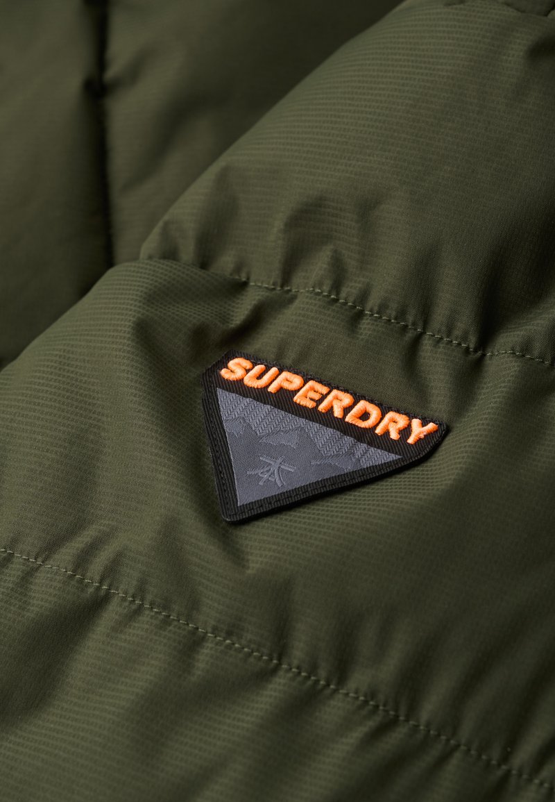 Tessuto imbottito verde oliva con texture trapuntata; presenta una toppa triangolare nera con "SUPERDRY" in un motivo arancione e grigio.