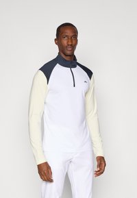 Camisa desportiva de mangas longas com corpo branco, gola e detalhes em azul-marinho, e mangas amarelo-claro. Apresenta um fecho de correr no pescoço.