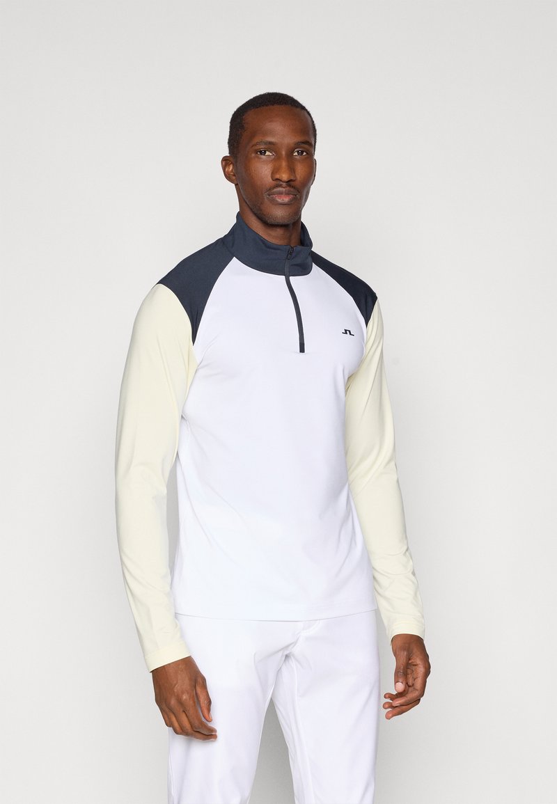 Camisa desportiva de mangas longas com corpo branco, gola e detalhes em azul-marinho, e mangas amarelo-claro. Apresenta um fecho de correr no pescoço.