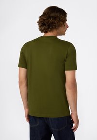 Man met krullend bruin haar, gekleed in een olijfgroen T-shirt met korte mouwen en donkerblauwe jeans, staand met de rug naar ons toe tegen een witte achtergrond.