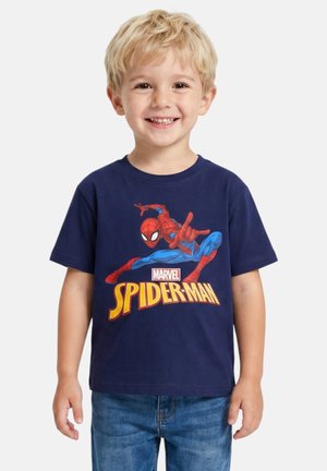 Jeune garçon blond souriant, portant un T-shirt bleu marine avec un graphique coloré de Spider-Man et un jean bleu sur un fond uni.