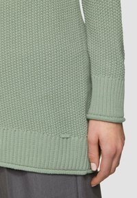 Hellgrüner Pullover mit einem strukturierten Strickmuster, gerippten Bündchen und Saum. Kleines Etikett mit "QS" am unteren Rand.