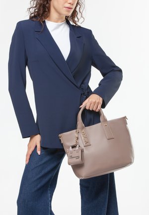 Sac à main en cuir beige avec des poignées doubles, des accessoires dorés et un charm amovible. Porté avec un blazer bleu marine et un pantalon en denim bleu.