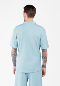 Hombre con los brazos tatuados, vistiendo una camisa de manga corta azul claro y pantalones cortos a juego, de pie de espaldas contra un fondo sencillo.