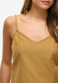 Camiseta mostaza con tirantes finos, escote en V y una textura suave. El tejido parece suave con detalles de costura mínimos.