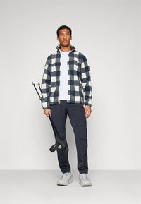 Lahe sinise ja kreemika ruudumustriga fliisjope, millel on esiosa lukk ja kõrge krae. Koos tumedate pükste ja hallide matkajalatsitega, käes matkakepid.